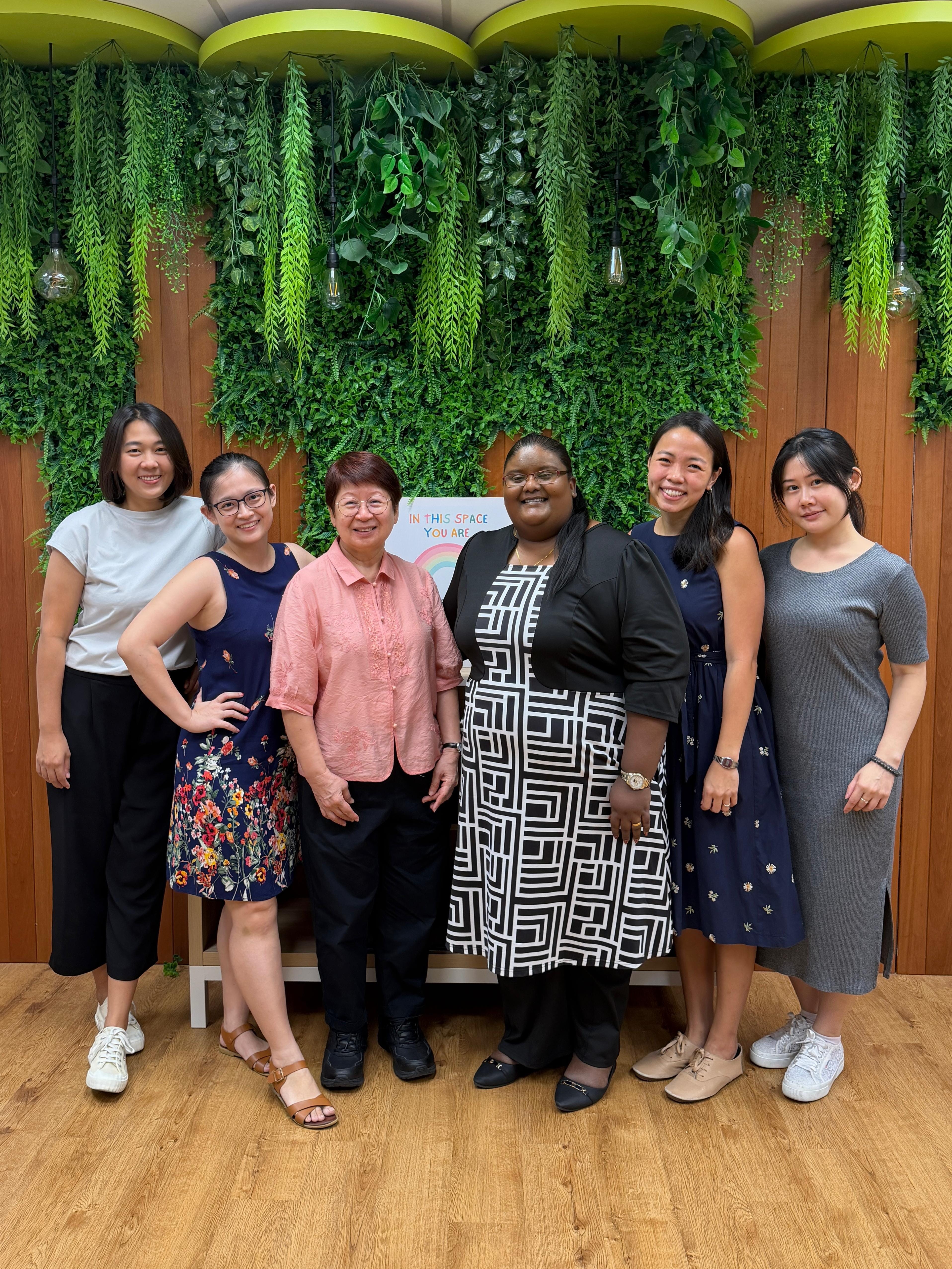 Six female counsellors posing together in front of a lush green plant wall, with a sign reading 'In This Space You Are...' visible in the background.